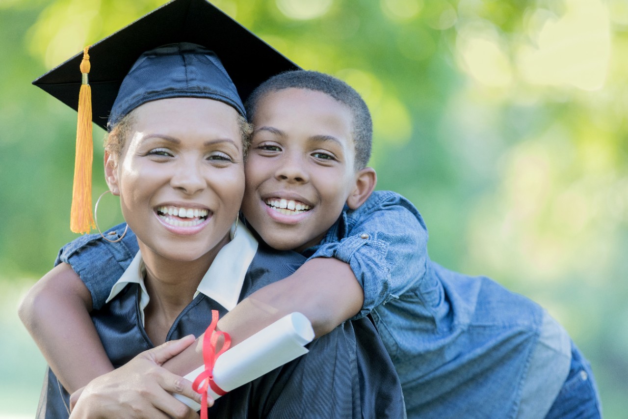 An adorable tween son stands outdoors behind his seated mother and drapes his arms over her shoulders.  They are cheek to cheek as they smile for the camera.  She wears a cap and gown and holds a rolled degree after her graduation ceremony.