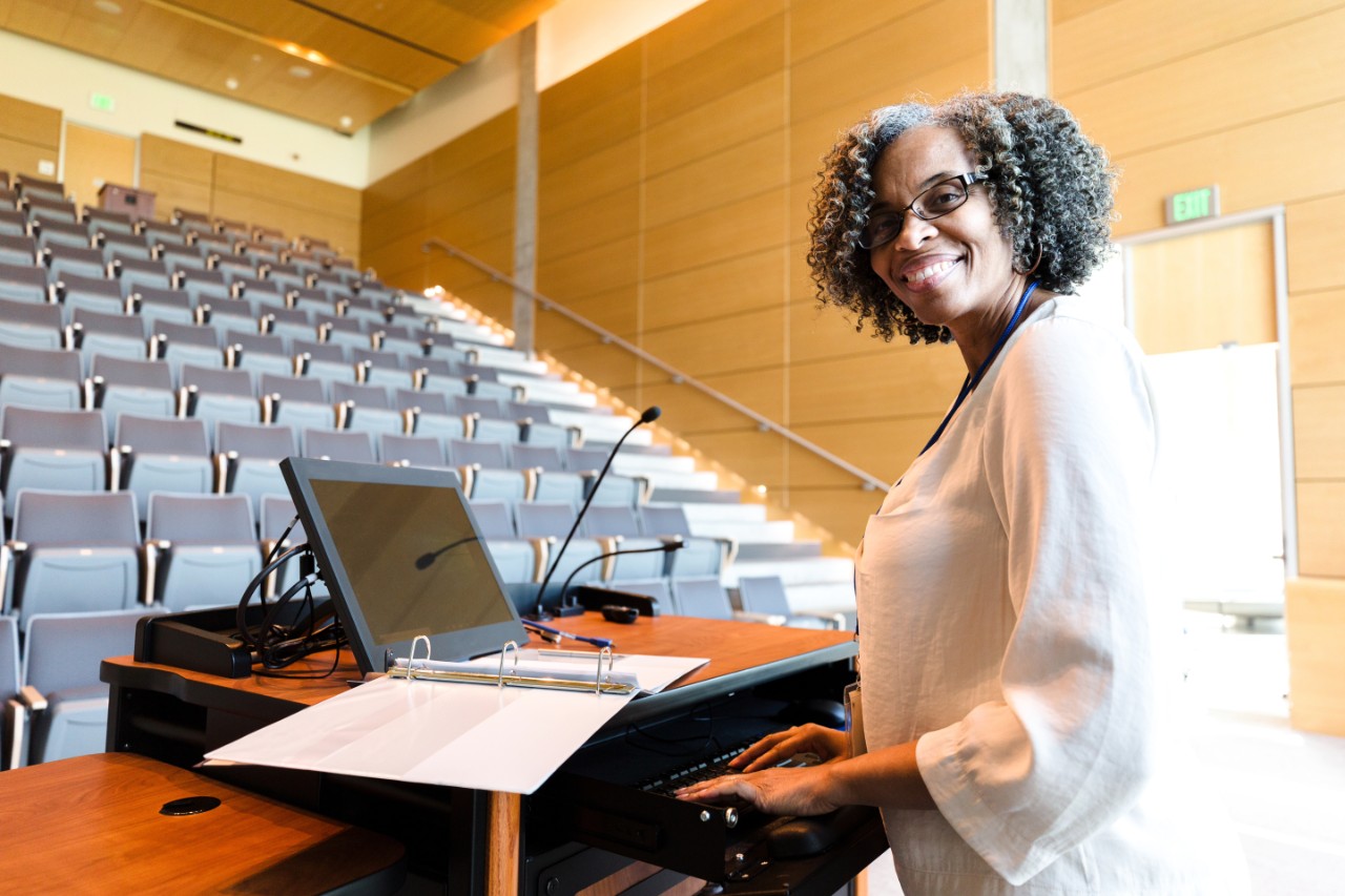 The cheerful, mature adult female university professor pauses in her class preparation to smile for the camera.  She is using the audio visual equipment.