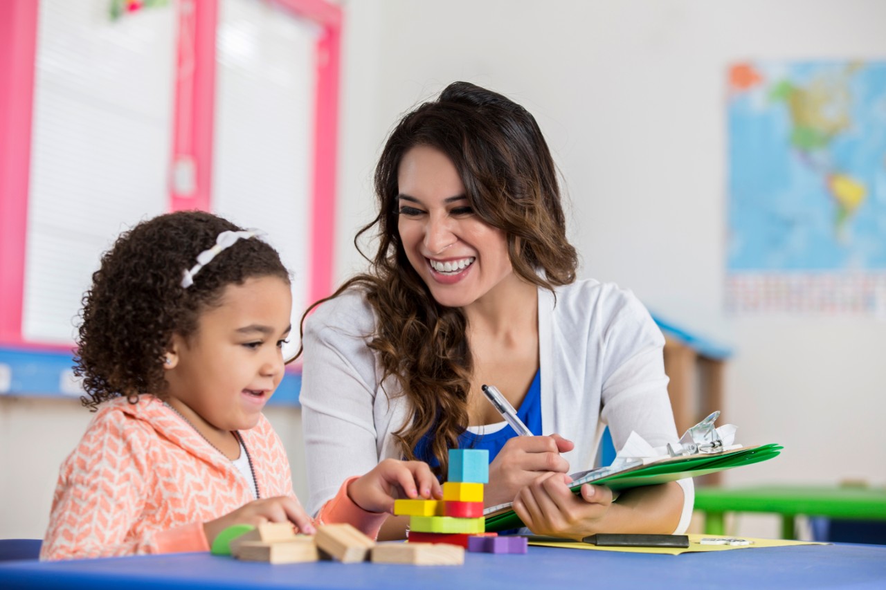 Smiling Hispanic young adult female teacher working with adorable African American little girl during preschool class. They are sitting at a table with colorful blocks. The teacher takes notes as the little girl puts the blocks together.